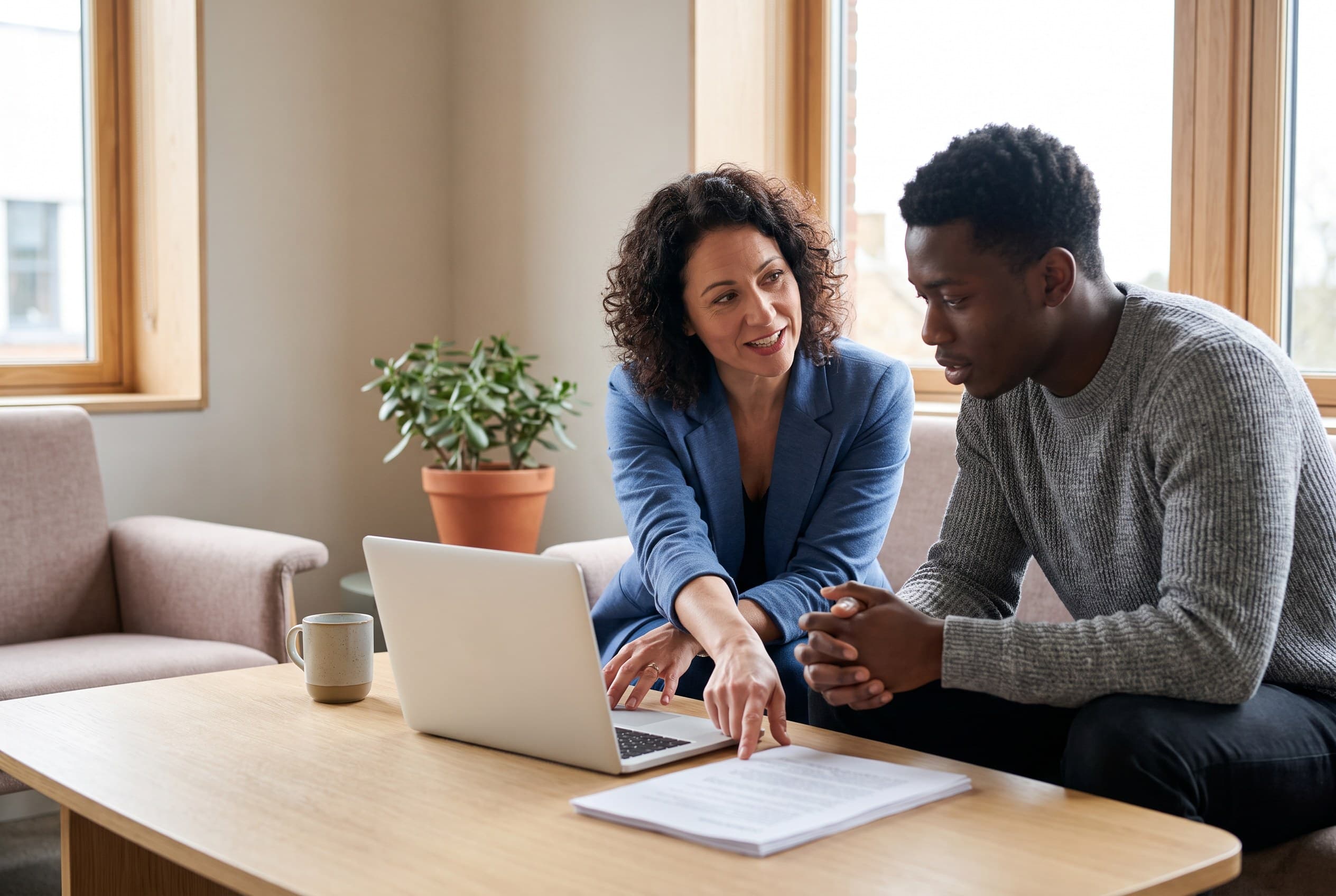 A career coach and a client sitting together at a coffee table, gesturing toward a laptop and a printed resume between them