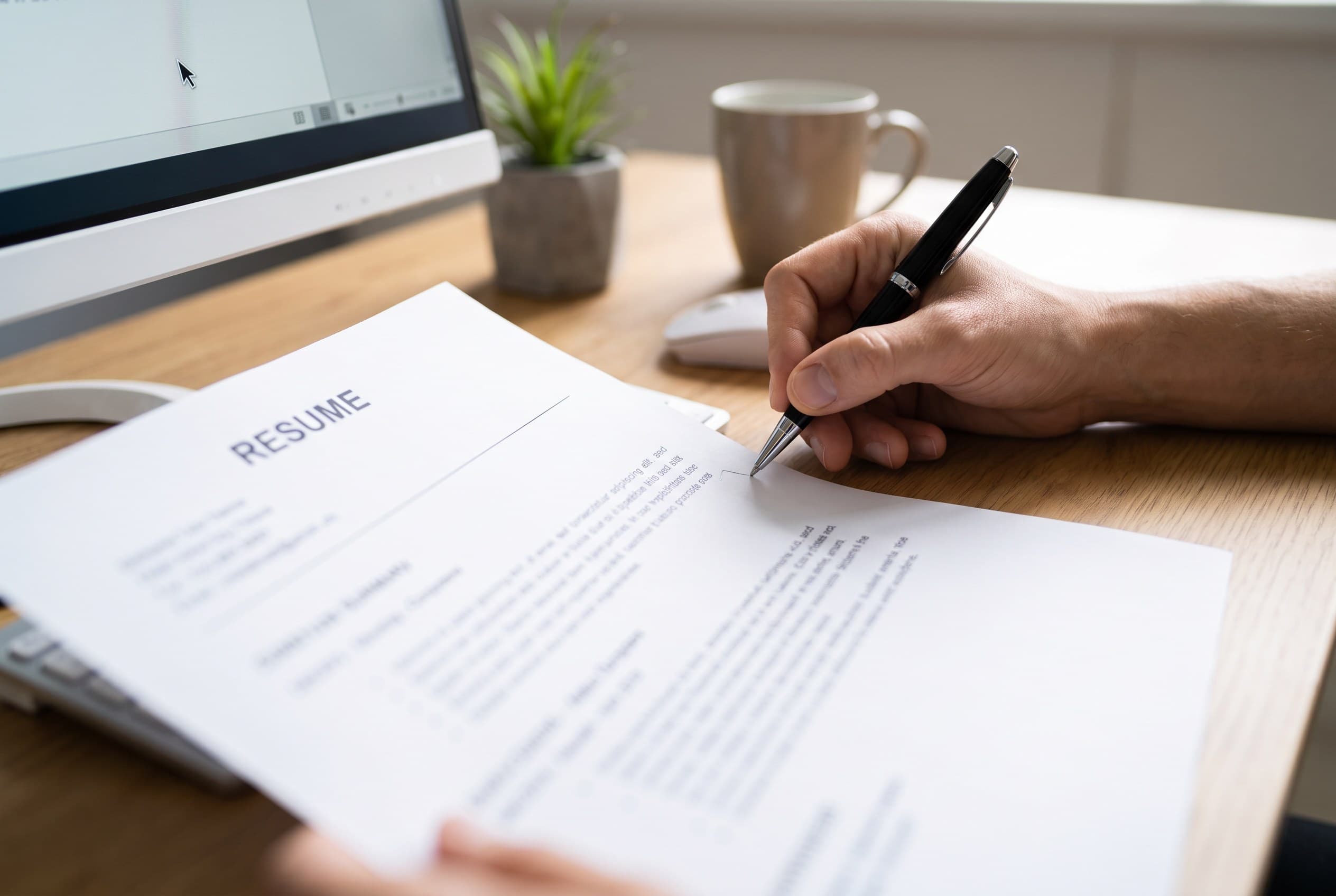 A hand marking up a printed resume with a pen beside a computer screen